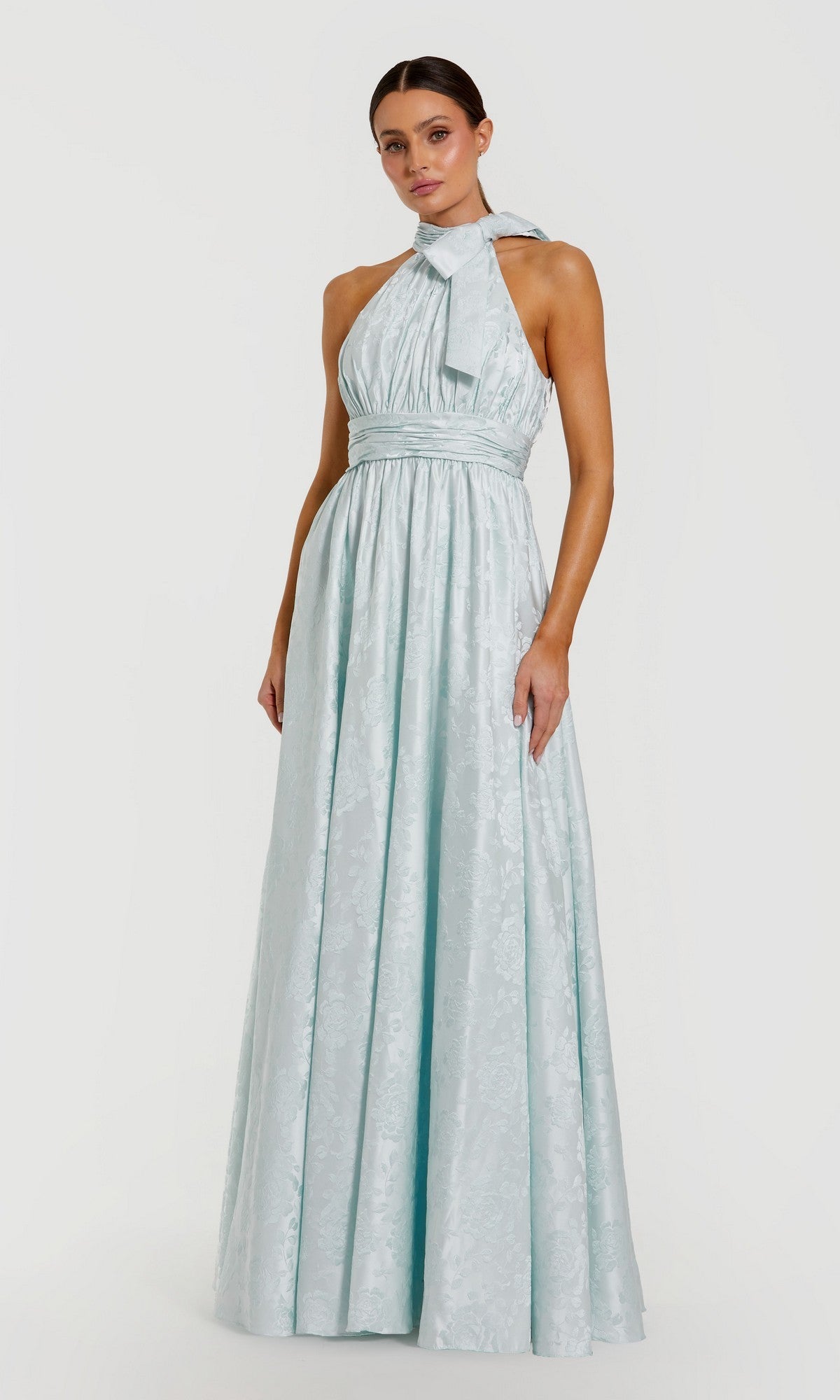 A woman stands against a plain white background wearing the Mac Duggal 56246 Long Formal Dress in powder blue, a halter-neck, floor-length gown featuring a bow detail at the neck.